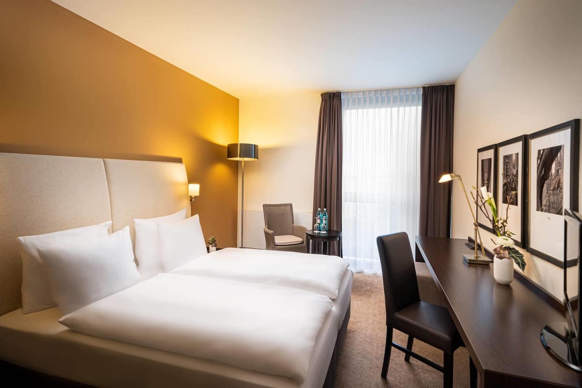 A hotel room with a large bed, a desk, a chair, a lamp, and a window with curtains.