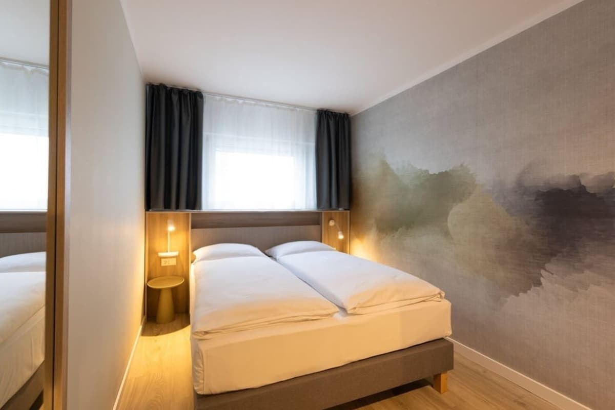 A hotel room with a bed, bedside lamps, a window with curtains, and a textured wall.