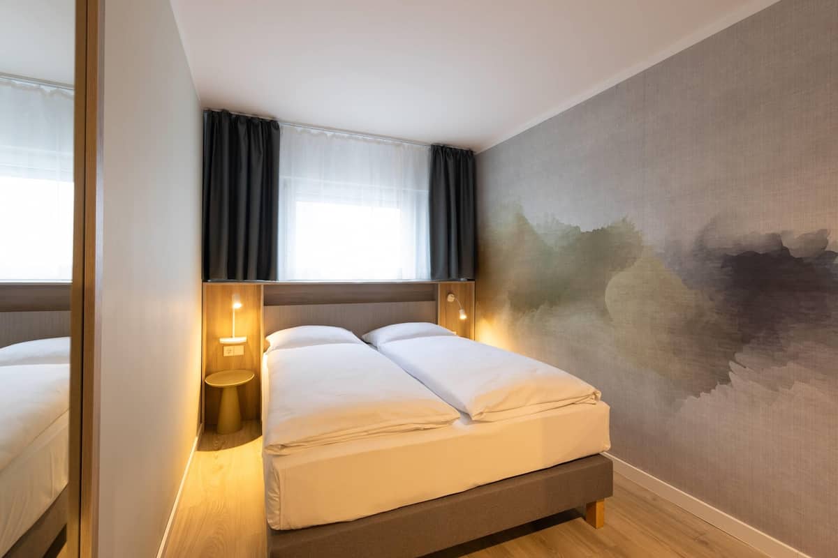 A hotel room with a bed, bedside lamps, a window with curtains, and a textured wall.