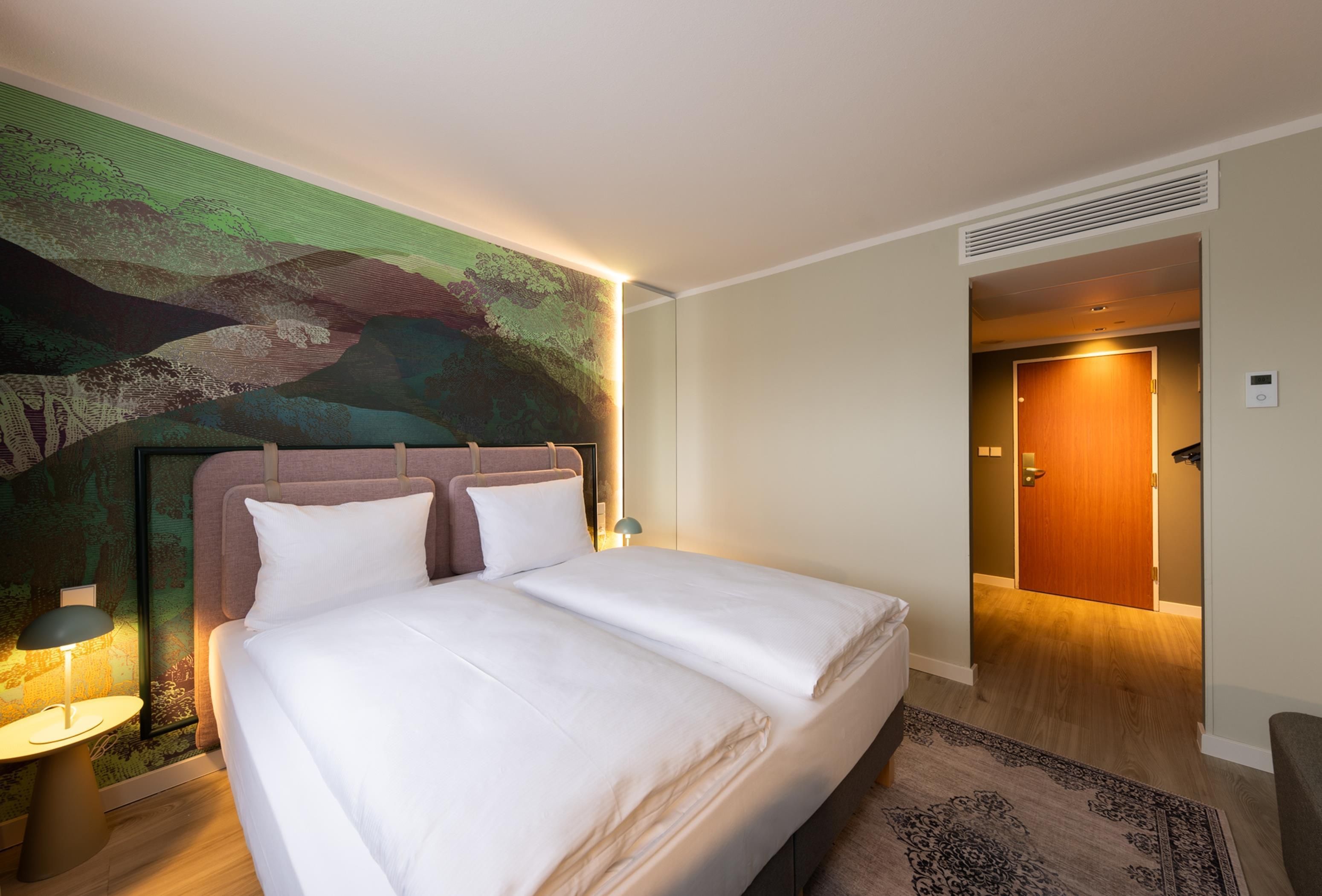Habitació d'hotel amb un llit, un capçal de llit, una obra d'art a la paret, una tauleta de nit, una làmpada i una porta que condueix a una altra habitació.