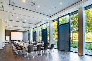 A conference room with a long table, chairs, a projector screen, and large windows with a view of trees. - The Rilano Hotel Munich, Trademark Collection by Wyndham (Munich)