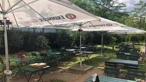 An outdoor dining area with green tables and chairs, covered by large umbrellas with logos, set in a garden with trees and a clear sky. - The Rilano Hotel Munich, Trademark Collection by Wyndham (Munich)
