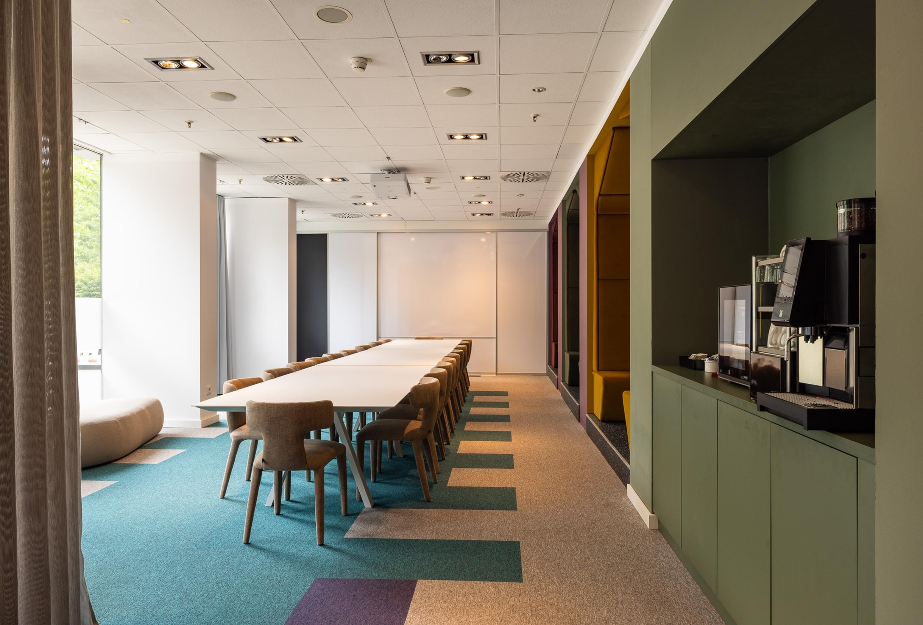 Une salle de conférence avec une longue table et des chaises, un appareil à café et une vue à travers une paroi en verre.