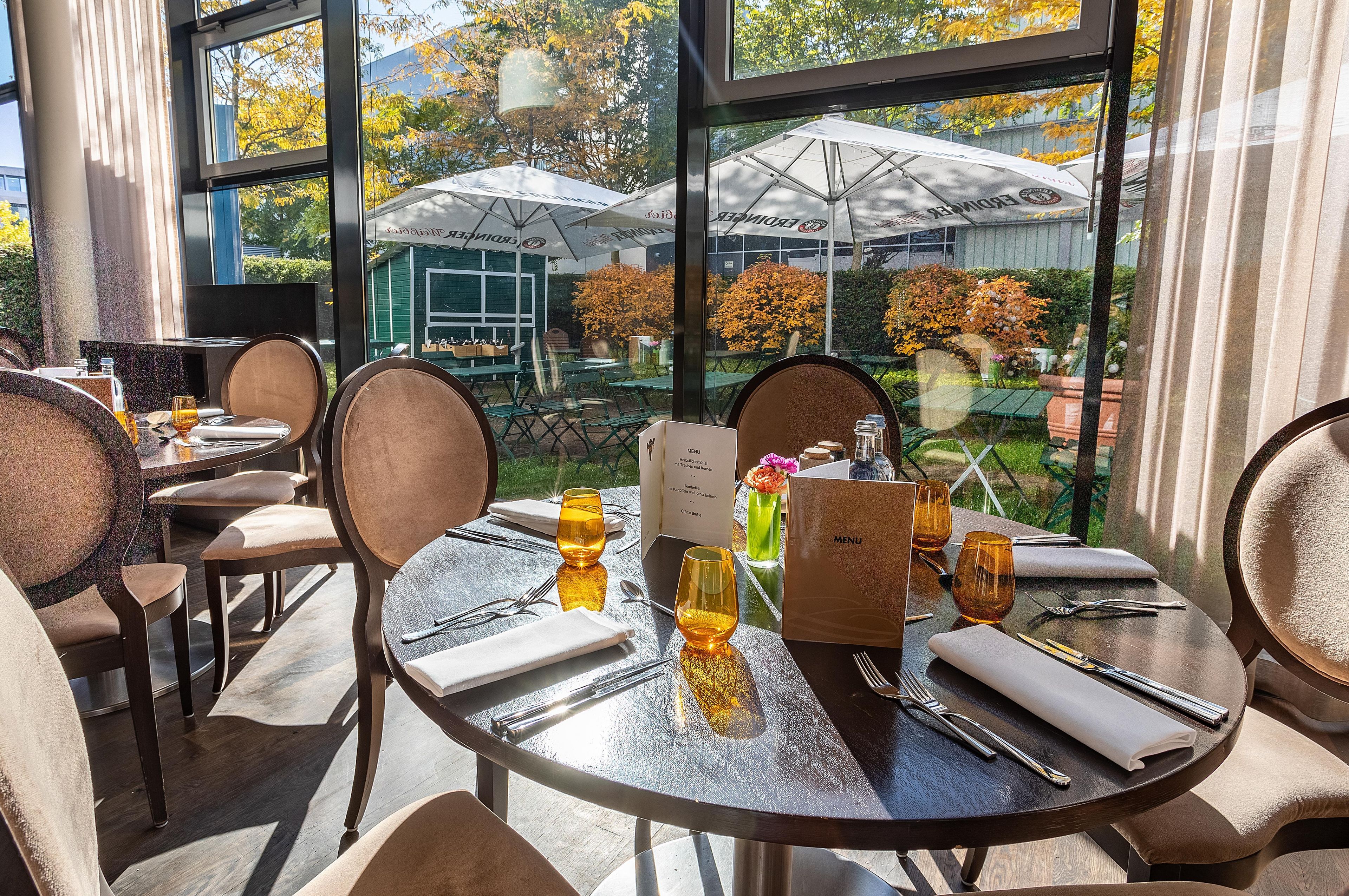 Un espace repas avec des tables rondes, des chaises et un présentoir à menus. À l’extérieur, on trouve des parasols et des arbres aux feuilles d’automne.