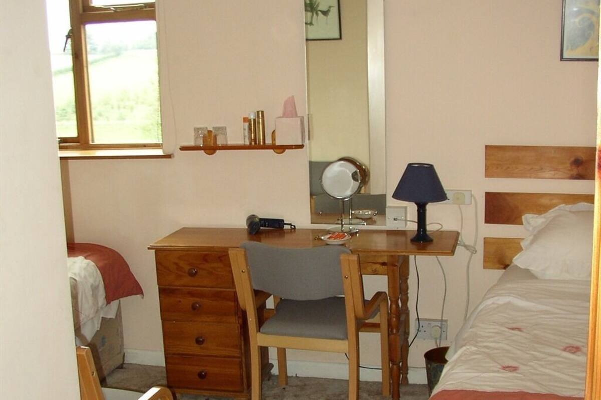 A bedroom with a wooden desk, chair, and a bed with a headboard. There is a mirror above the desk and a lamp on it.