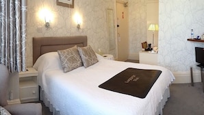 Una cama bien hecha con edredón blanco y dos almohadas, una mesita de noche con lámpara, y un escritorio pequeño con ordenador.