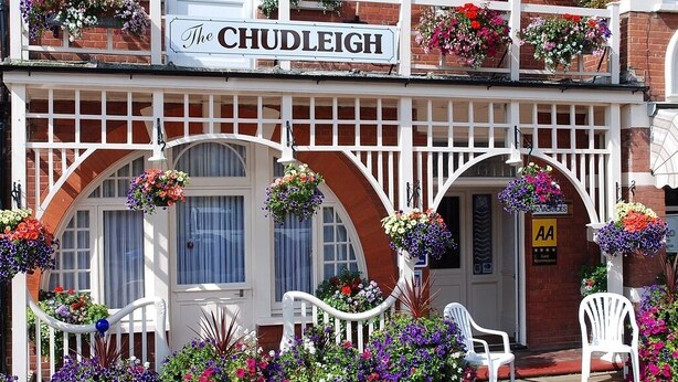 Un pub tradicional llamado 'The CHUDLEIGH' con fachada blanca, ventanas arqueadas y macetas colgantes de flores.