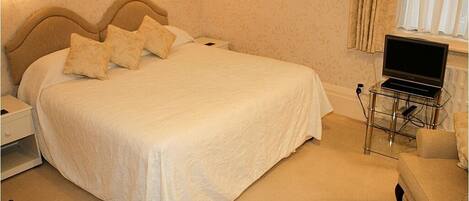 A neatly made bed with a white bedspread and pillows, a bedside table with a lamp, a laptop on a stand, and a chair in a hotel room.