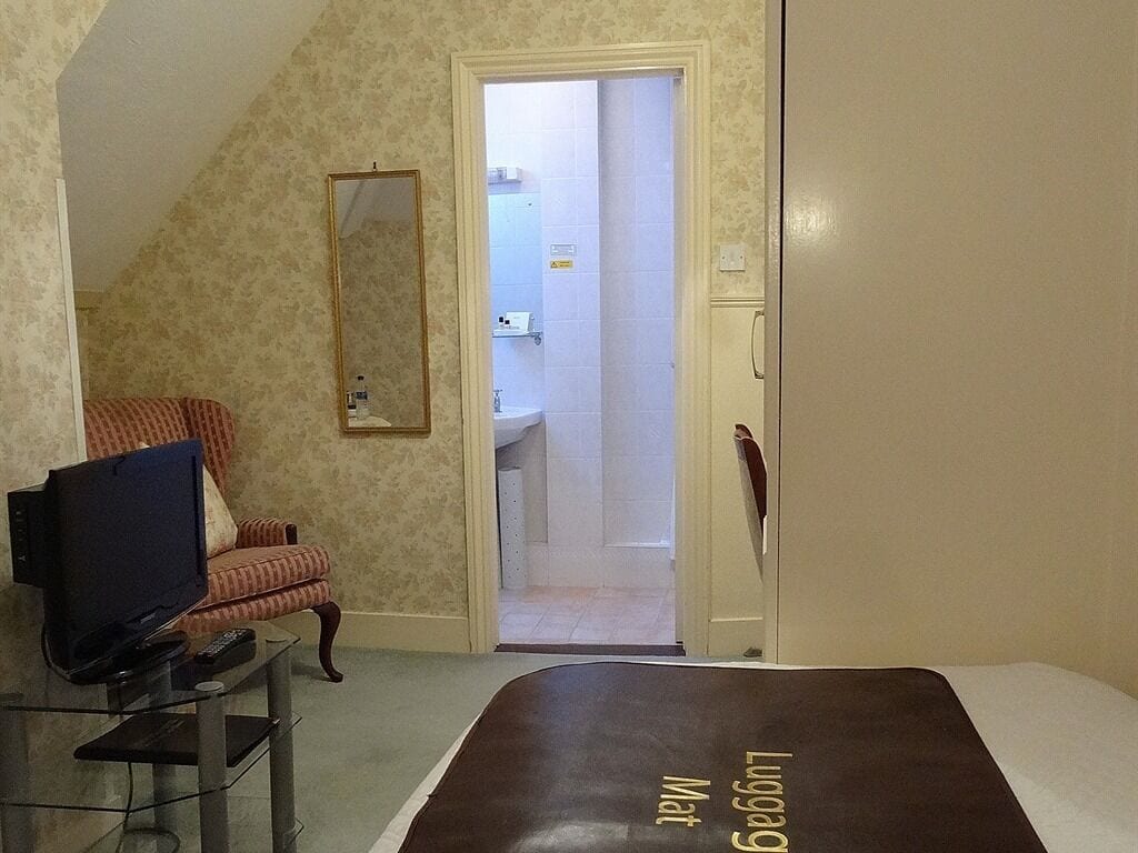 A hotel room with a bed, a television on a glass table, a chair, and a bathroom visible through an open door.