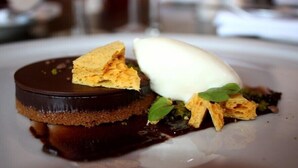 Postre amb base de xocolata, una bola de gelat i elements cruixents damunt d'un plat.