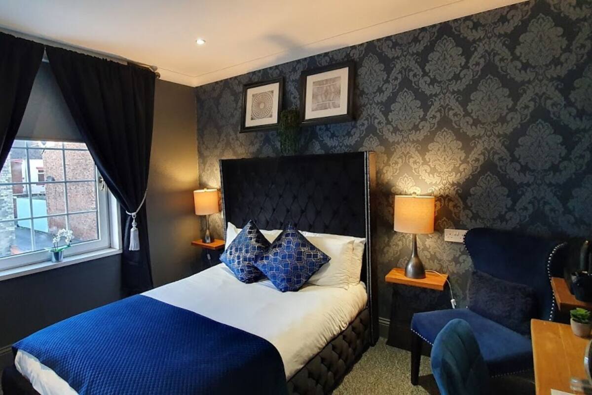 Habitación de hotel con una cama, dos mesitas de noche, una silla, una ventana con cortinas y papel tapiz estampado.