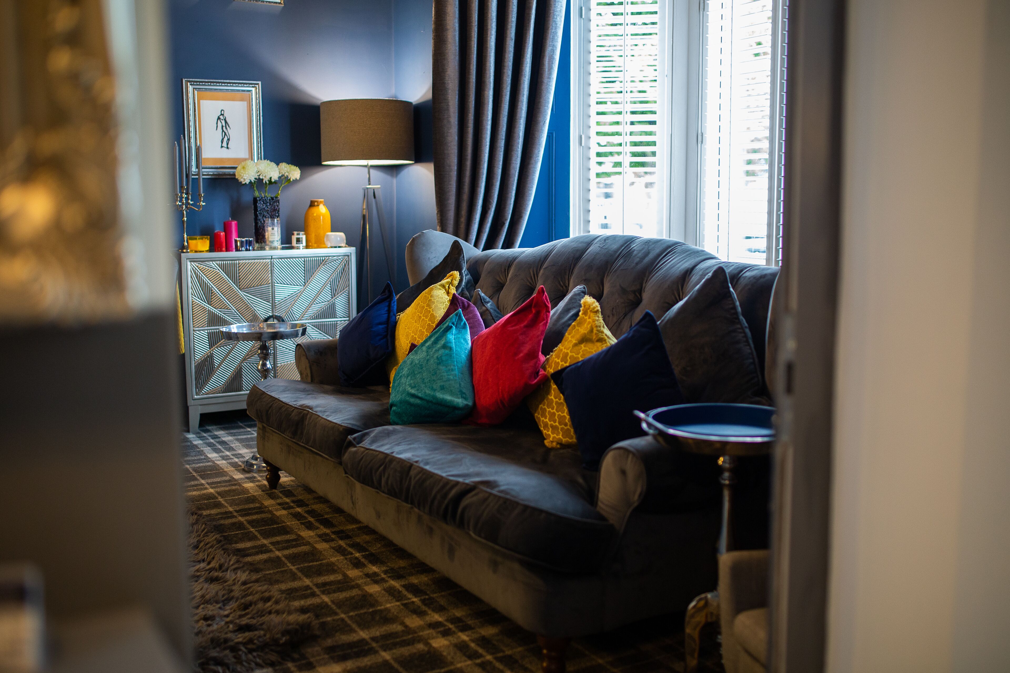 A living room with a dark-colored sofa, colorful cushions, a patterned rug, a side table with decorative items, and a window with blinds.