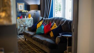 A living room with a dark-colored sofa, colorful cushions, a patterned rug, a side table with decorative items, and a window with blinds.