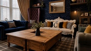 Ein Zimmer mit einem großen Holztisch, zwei Sofas mit gemusterten Kissen, einer dunkelblauen Wand mit Muster, einem Spiegel und einem Globus auf einem Regal.