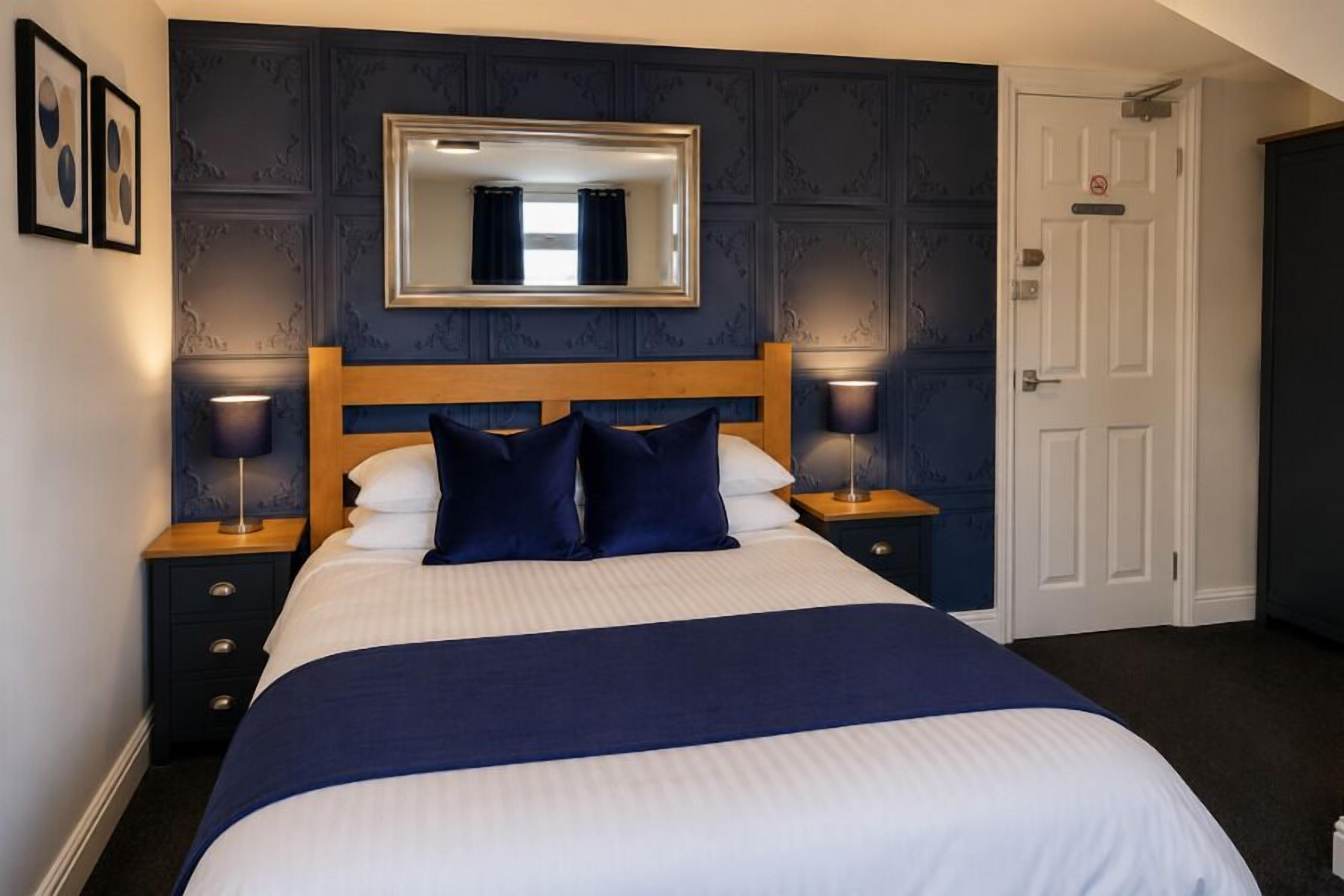 A neatly made bed with a wooden headboard, two bedside tables with lamps, a mirror above the bed, and a door to the right.