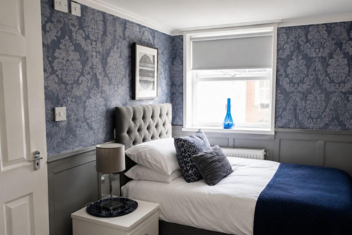 A bedroom with a tufted headboard, a bedside table with a lamp, a window with a view, and patterned wallpaper.