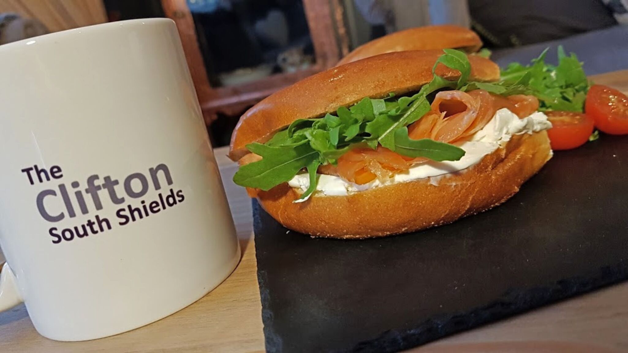 Sanduíche com rúcula e salmão defumado no pão, acompanhado de uma caneca branca com a inscrição 'The Clifton South Shields'.