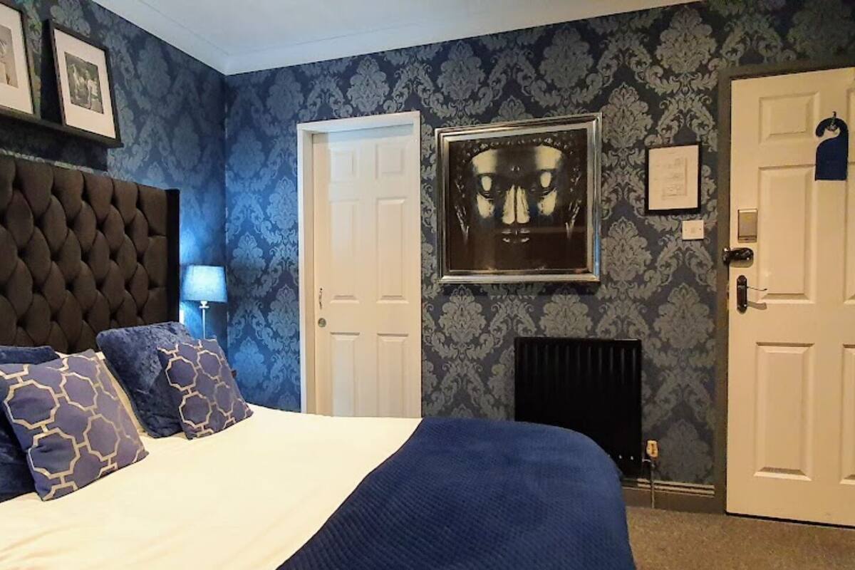 Un dormitorio con cabecera capitonada, cama con ropa de cama azul y blanca, un cuadro enmarcado en la pared y una puerta con un letrero.