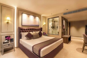 A hotel room with a large bed, bedside lamps, a painting, a mirror, a small table, and a chair.