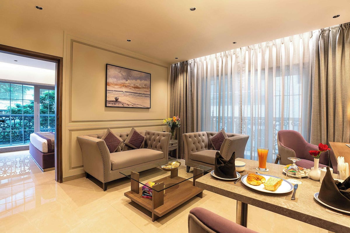 Una habitación de hotel moderna con sofá, sillones, una mesa de comedor con desayuno y un balcón con vistas.