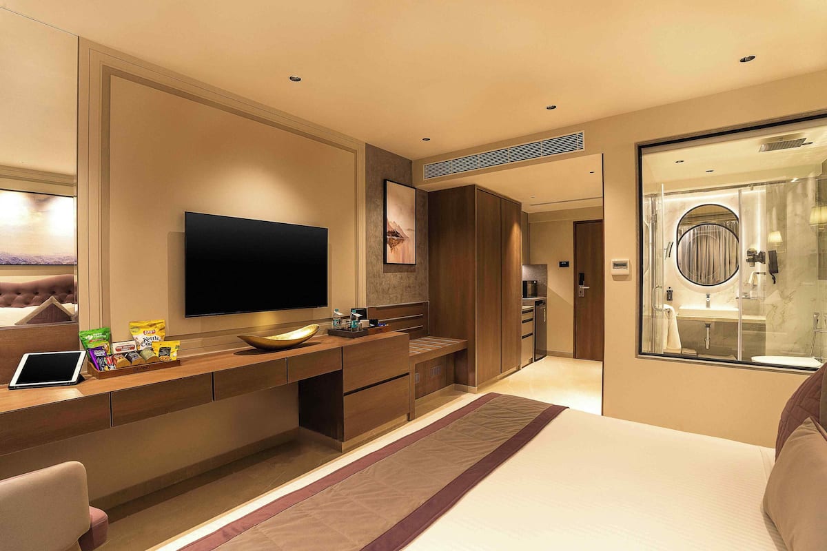 Una habitación de hotel moderna con un televisor de pantalla plana grande, una mesa consola de madera con aperitivos, una cama con cabecera y un baño con paredes de vidrio.