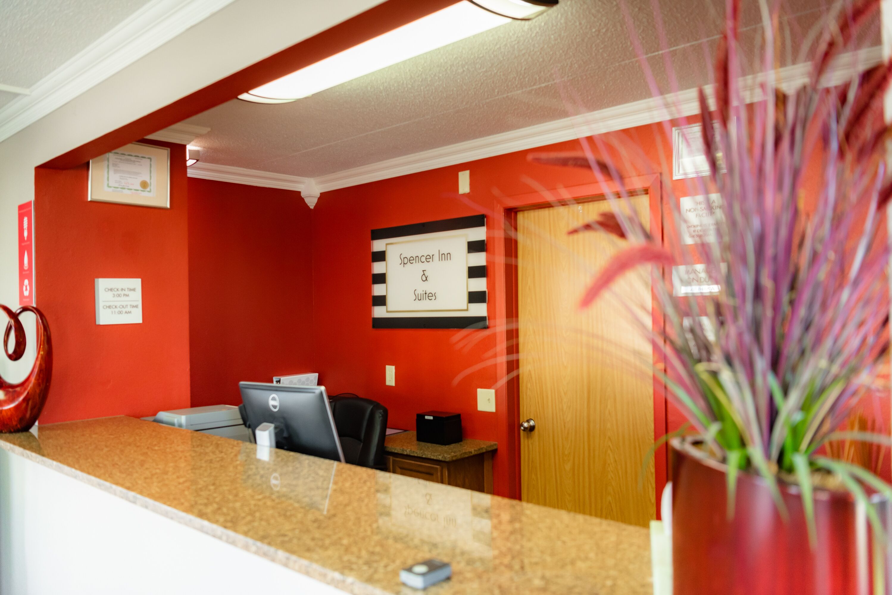 Uma área de recepção de hotel com parede vermelha, sinalização para Spencer Inn Suites, balcão de recepção, computador e uma planta.