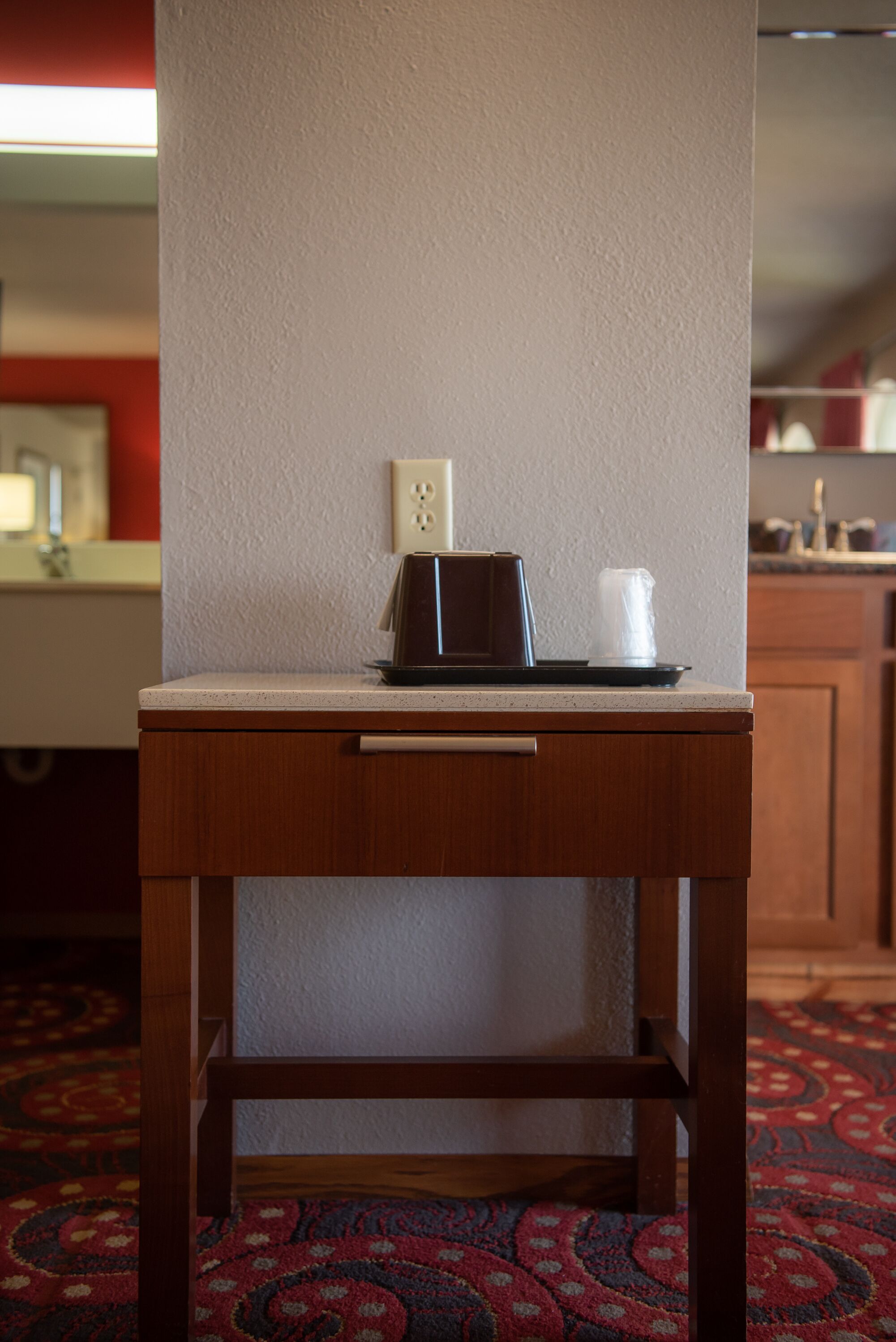 A small wooden table with a microwave and a cup on it, positioned against a wall with a power outlet.