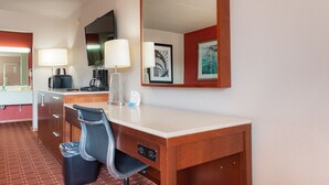 Ein Hotelzimmer mit Schreibtisch aus Holz, zwei Lampen, einem Sessel, einem Papierkorb, einer Kaffeemaschine, einem Fernseher und gerahmten Bildern an der Wand.