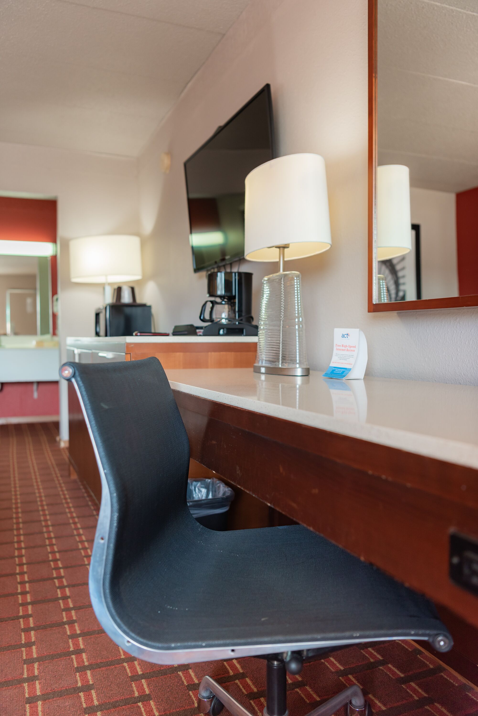 A hotel room with a desk, a chair, a lamp, a coffee maker, and a flat-screen TV.