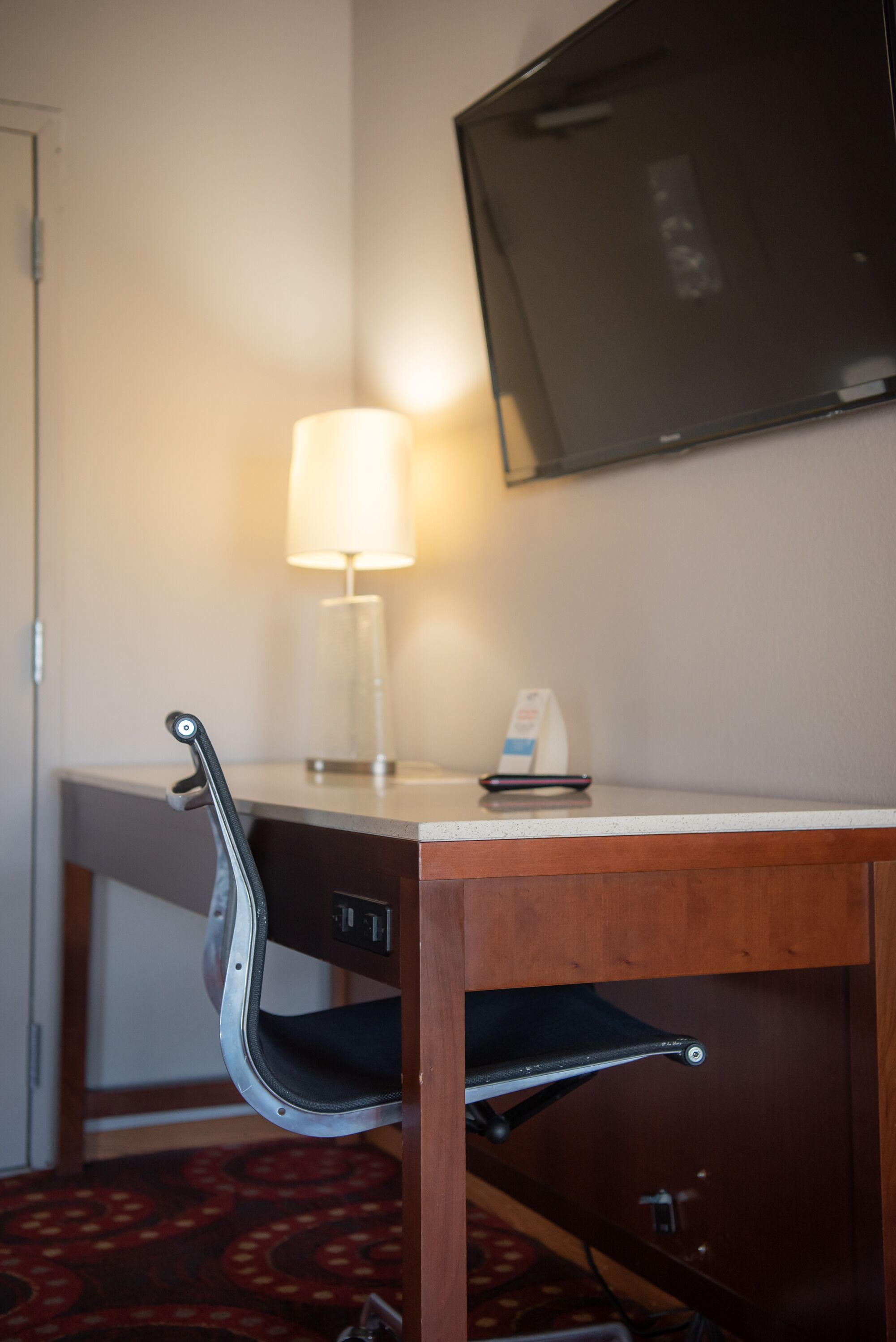 A hotel room with a wooden desk, a lamp, a television mounted on the wall, and a chair with a tray.