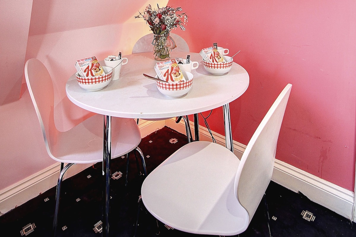 A room with a pink wall, a mounted television displaying 'Insight Radio', a white table with two chairs, and a vase with flowers.