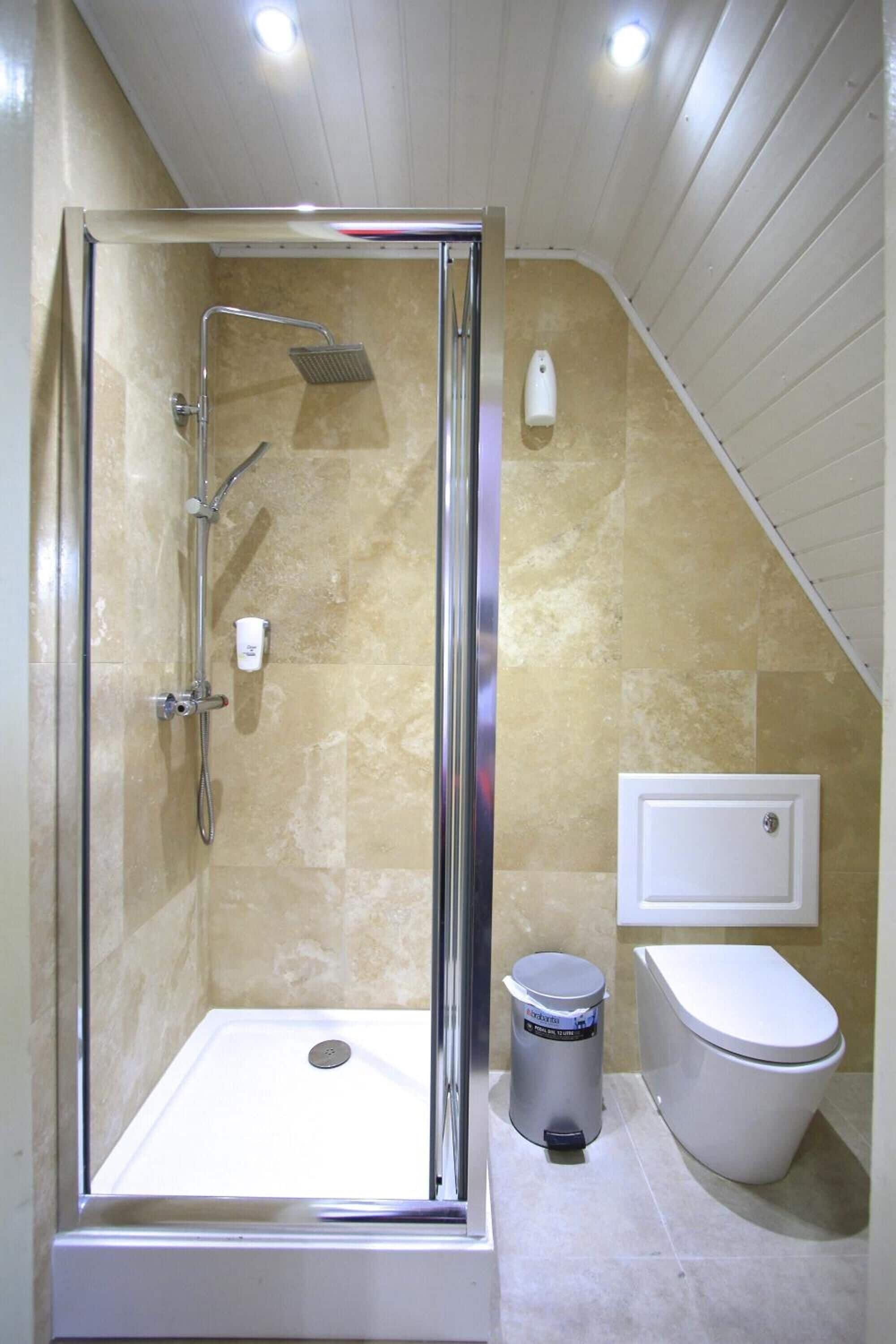 Ein Duschbereich mit Glastür, Handbrause, Wandseifenspender und Toilette mit einem Mülleimer daneben.