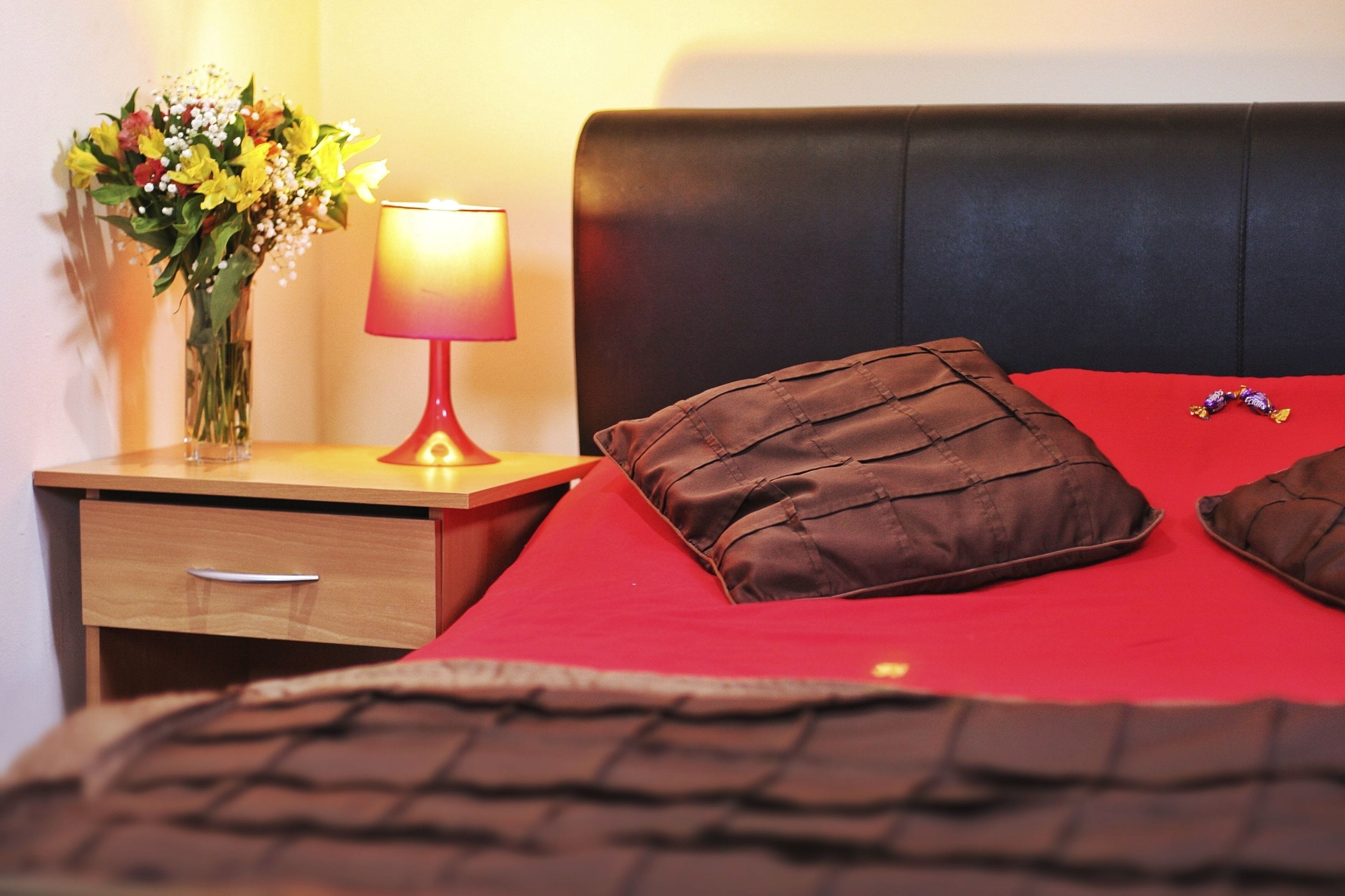 A neatly made bed with a red blanket and two brown pillows, a bedside table with a lamp and a vase of flowers, and a wooden nightstand with a drawer.