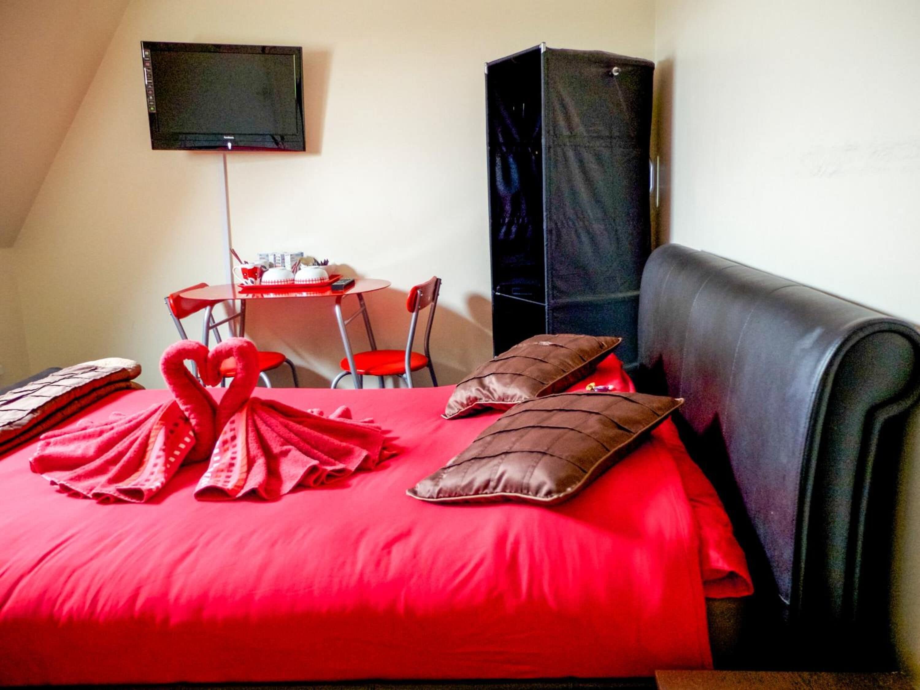 Quarto de hotel com colcha vermelha, TV de tela plana na parede, mesa pequena com cadeiras e um alto-falante preto.