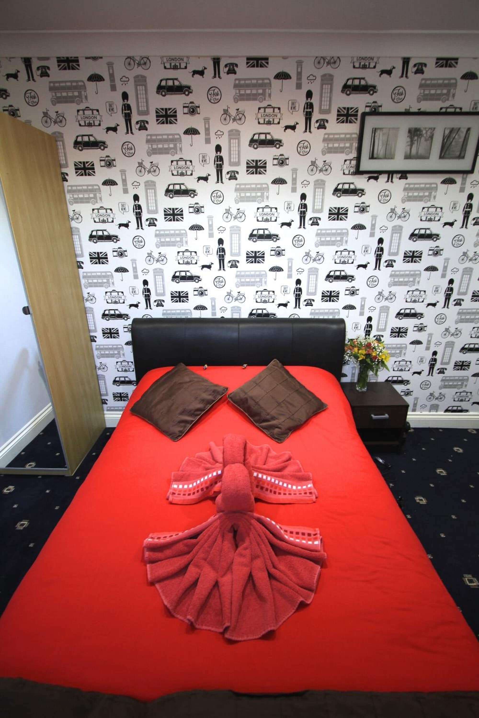 A hotel room with a red bedspread, two brown pillows, and a red towel arranged on the bed. The room features a patterned wallpaper with various icons and a framed picture on the wall.