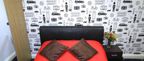 A hotel room with a red bedspread, two brown pillows, and a red towel arranged on the bed. The room features a patterned wallpaper with various icons and a framed picture on the wall.