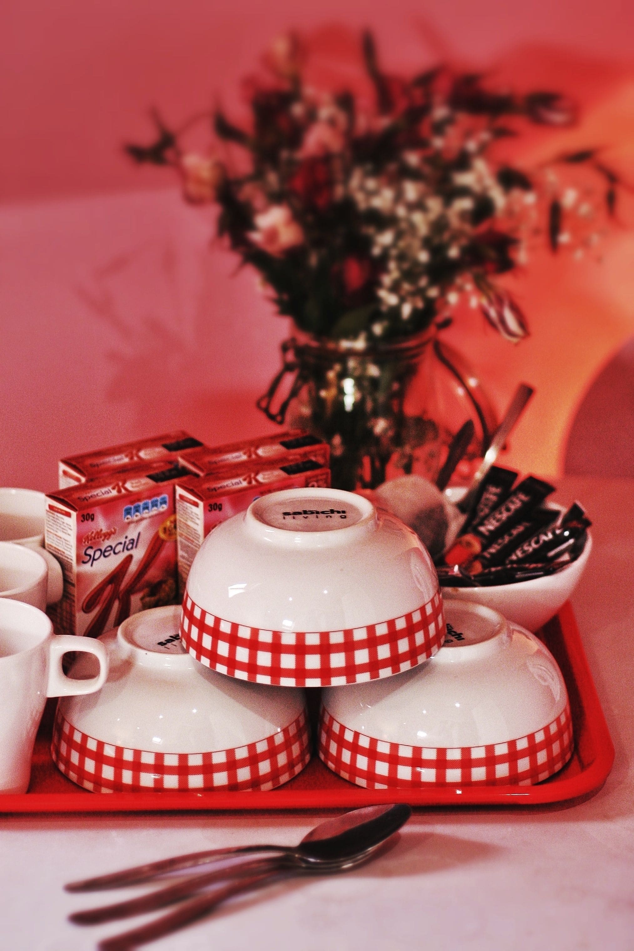 Uma bandeja com tigelas quadriculadas em branco e vermelho, uma caixa de café Nestlé Special, um vaso com flores e uma colher.