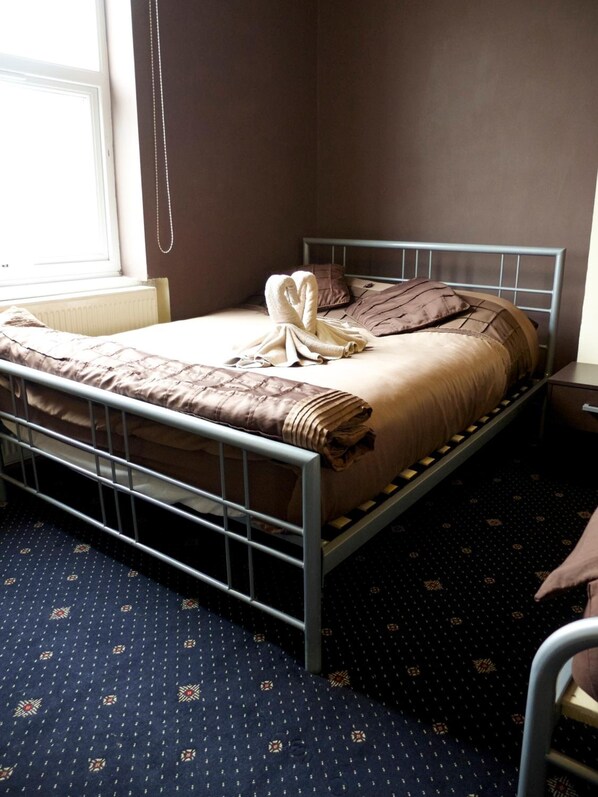 A single bed with a metal frame, a patterned carpet, and a window with blinds.