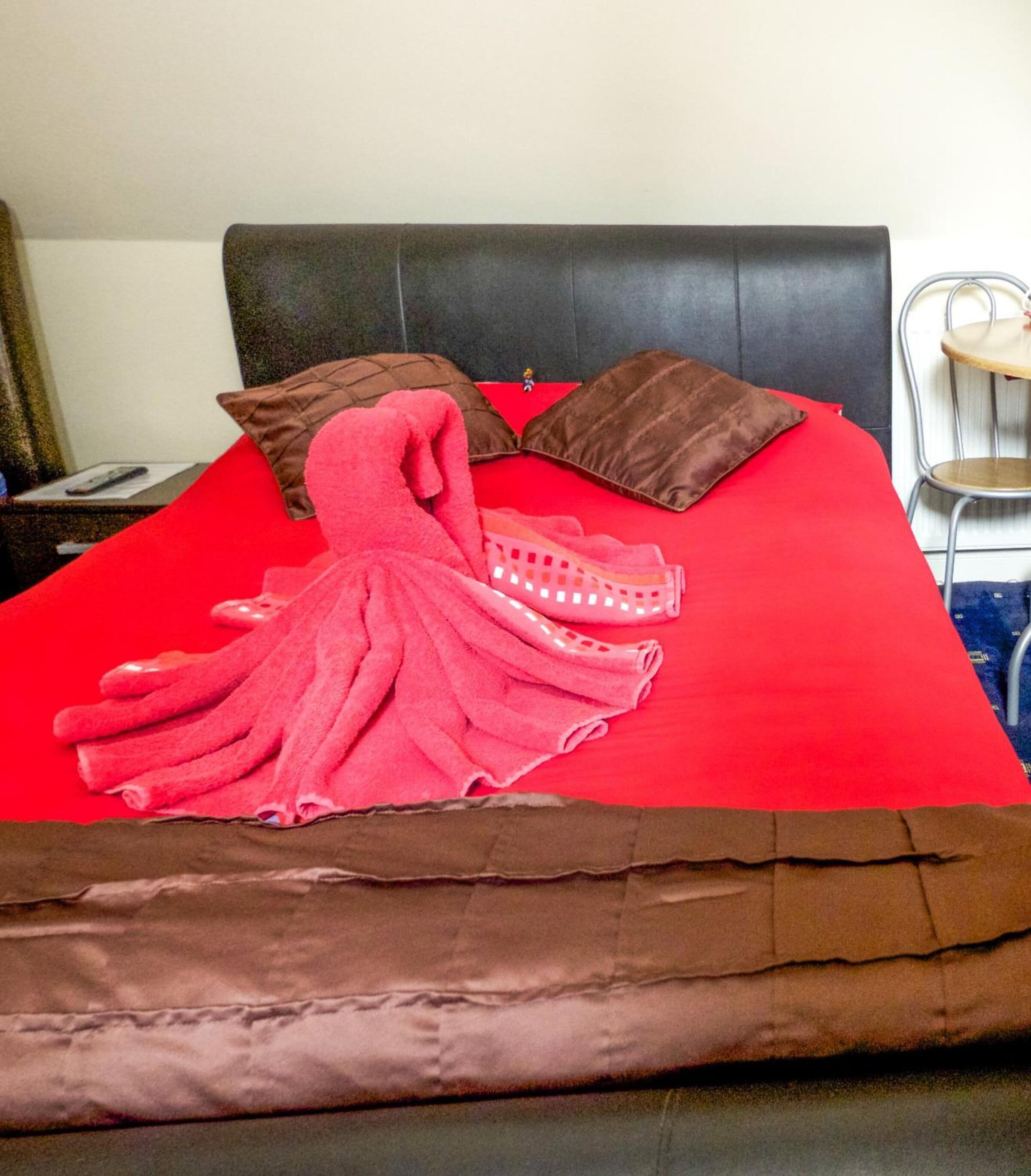 A bed with a red bedspread, two brown pillows, and a pink towel.