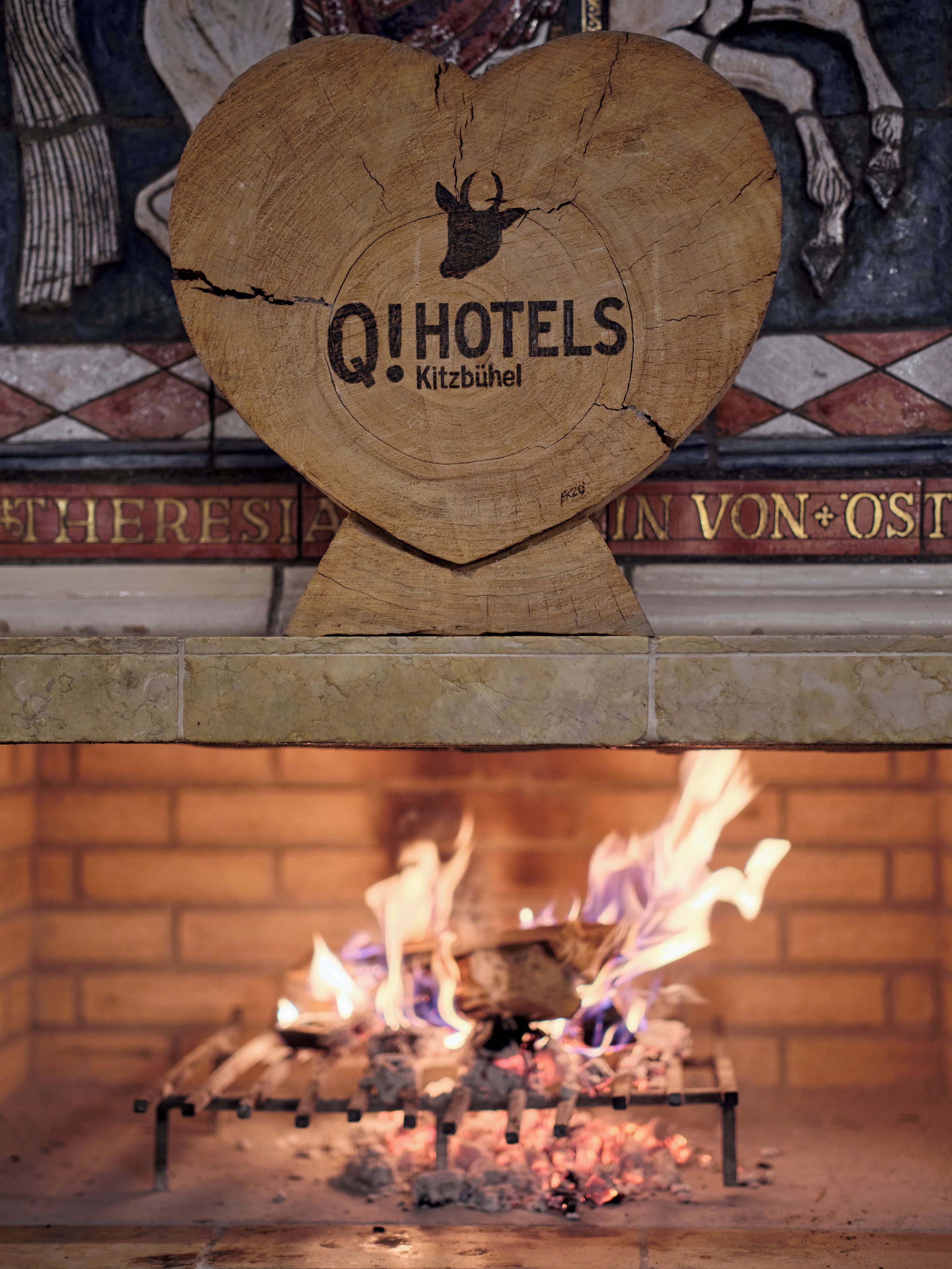 Una placa de madera con 'QIHOTELS' y 'Kitzbühel' grabados, montada sobre una chimenea con llamas visibles.