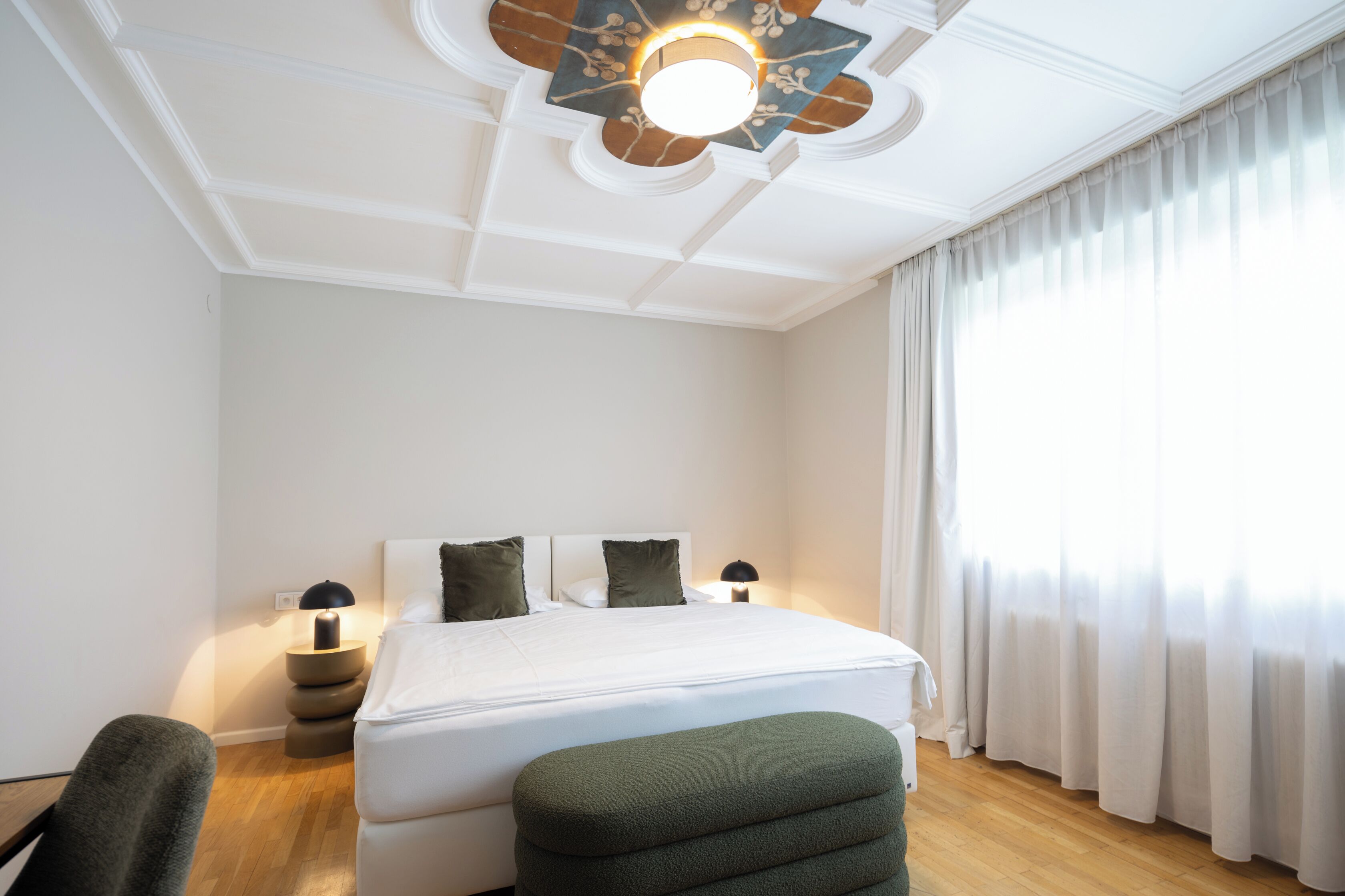 A hotel room with a bed, two bedside tables, a chair, a ceiling fan, and a window with curtains.