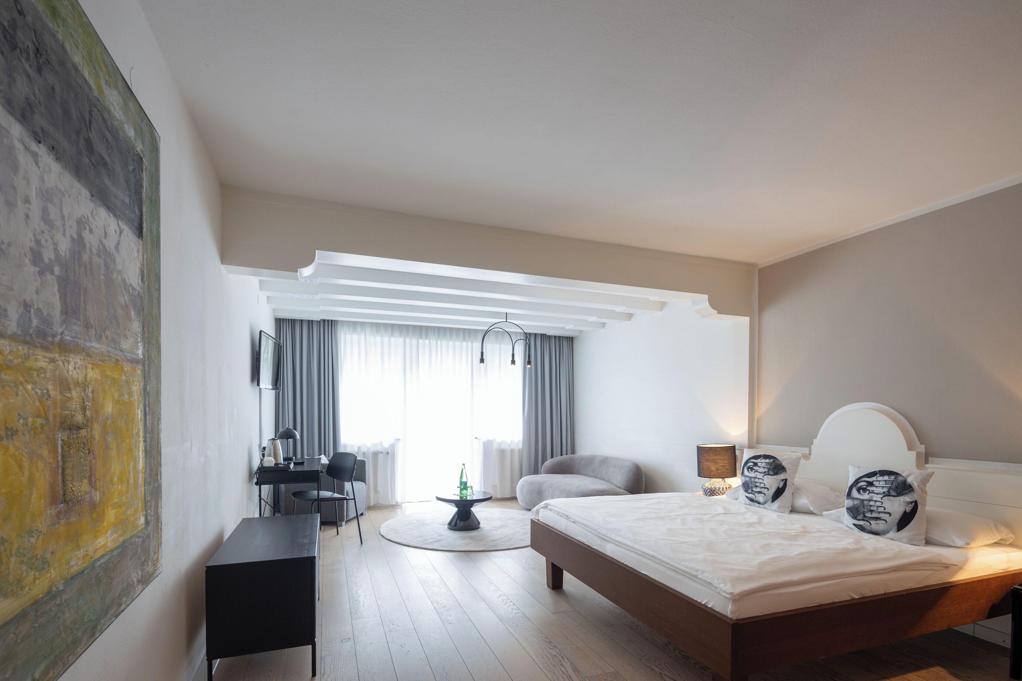 Quarto moderno com uma cama grande, uma mesa de trabalho e uma cadeira. Há uma janela com cortinas, uma mesa redonda e um quadro na parede.