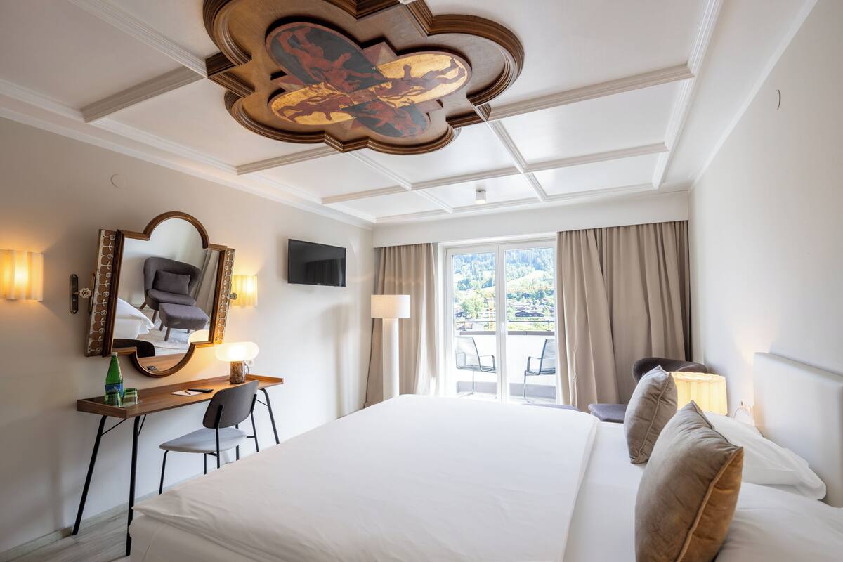A hotel room with a large bed, a desk with a chair, a mirror, a television, and a window with curtains.