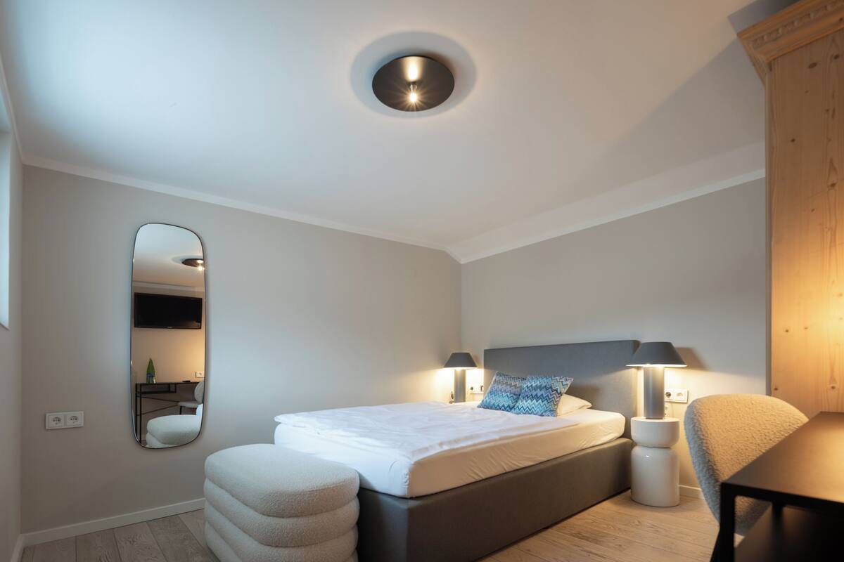 A modern bedroom with a bed, bedside lamps, a round ceiling light, a mirror, and a wooden wardrobe.