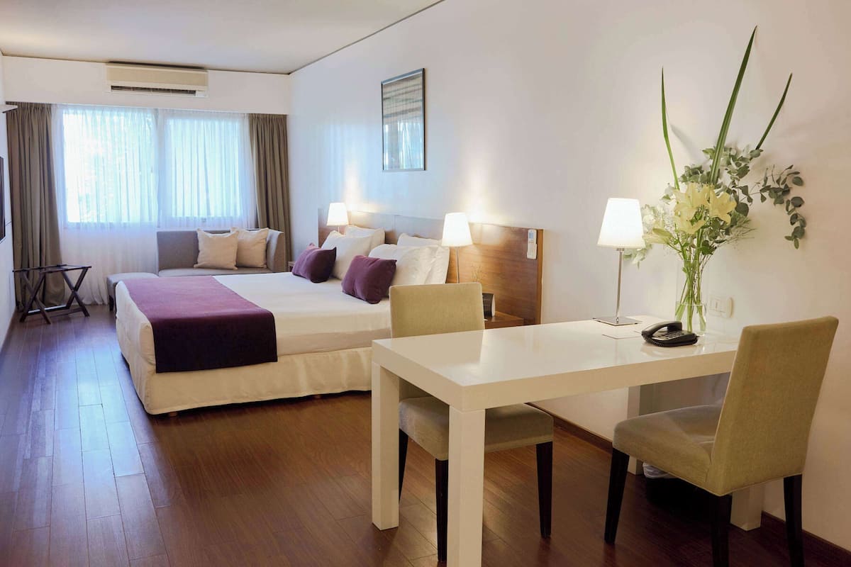 Ein Hotelzimmer mit zwei Betten, einem Schreibtisch und einem Stuhl. Es gibt ein Fenster mit Vorhängen, eine Wandlampe und eine Vase mit Blumen.