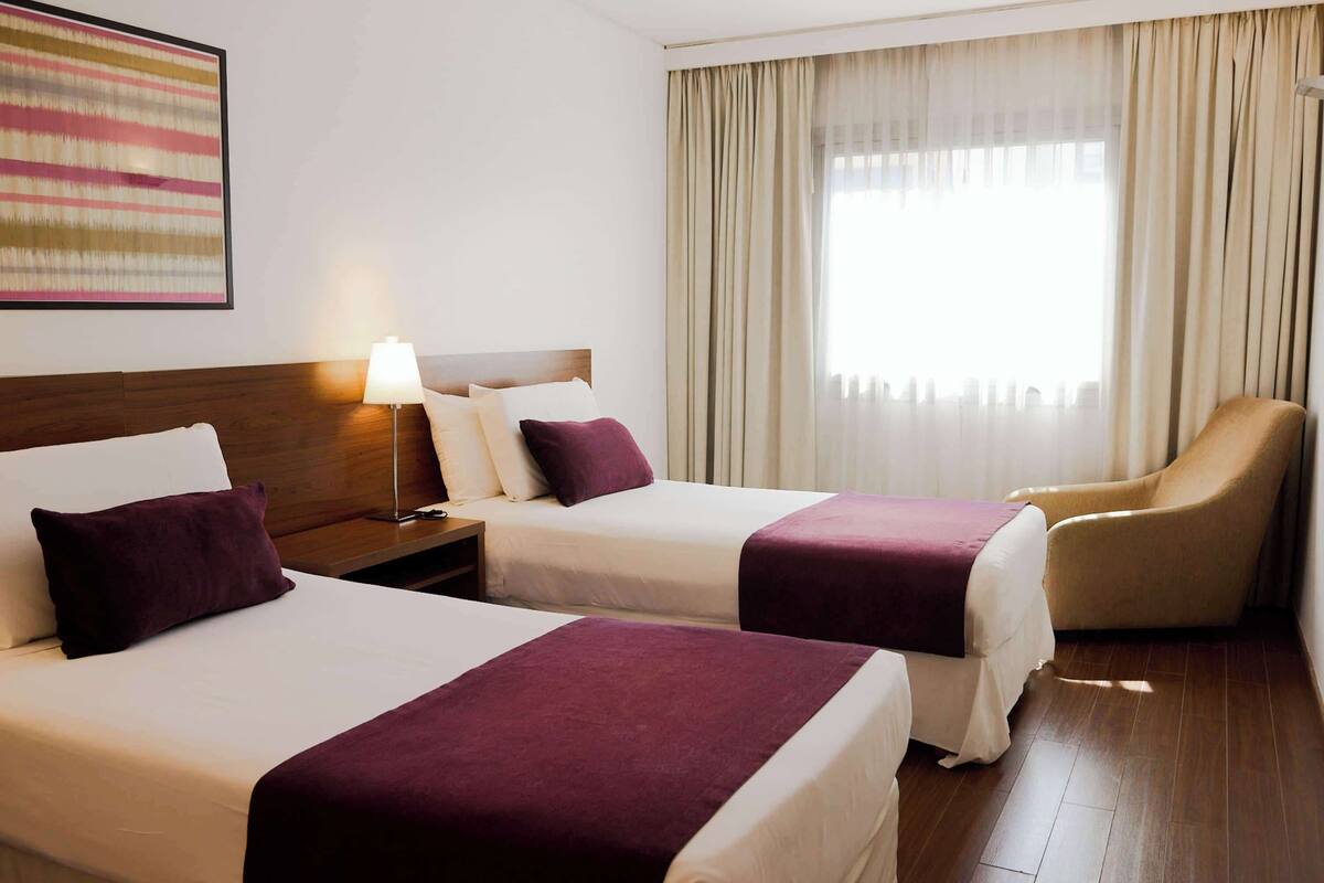 Ein Hotelzimmer mit zwei Betten, einem Sessel, einer Lampe und einem Fenster mit Vorhängen.