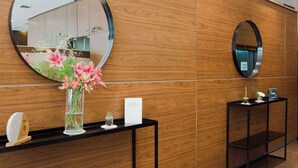 A reception area with a wooden wall, a round mirror, a black console table with a vase of flowers, and a small potted plant. - Dazzler by Wyndham Buenos Aires Maipu (Buenos Aires)