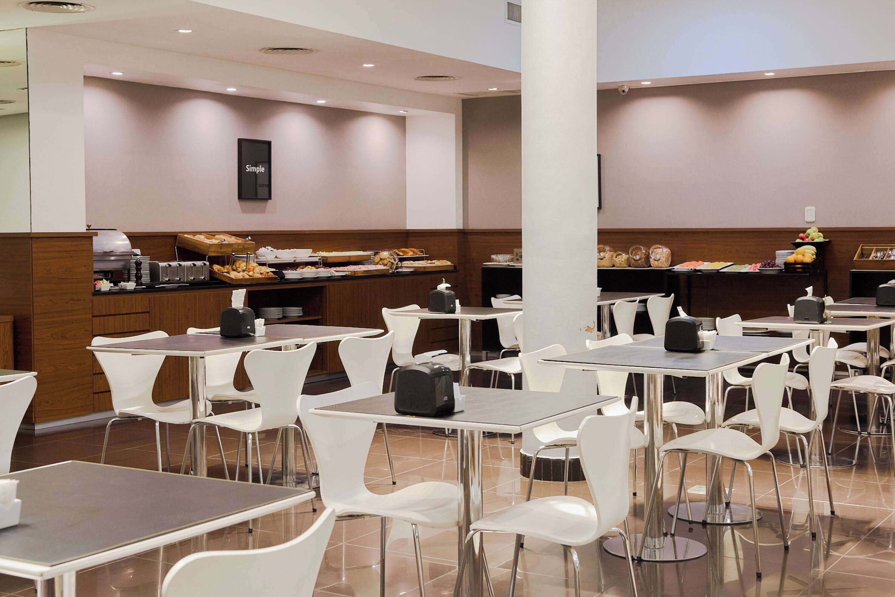 A modern dining area with white chairs and tables, a buffet with various food items, and a "Simple" sign on the wall.