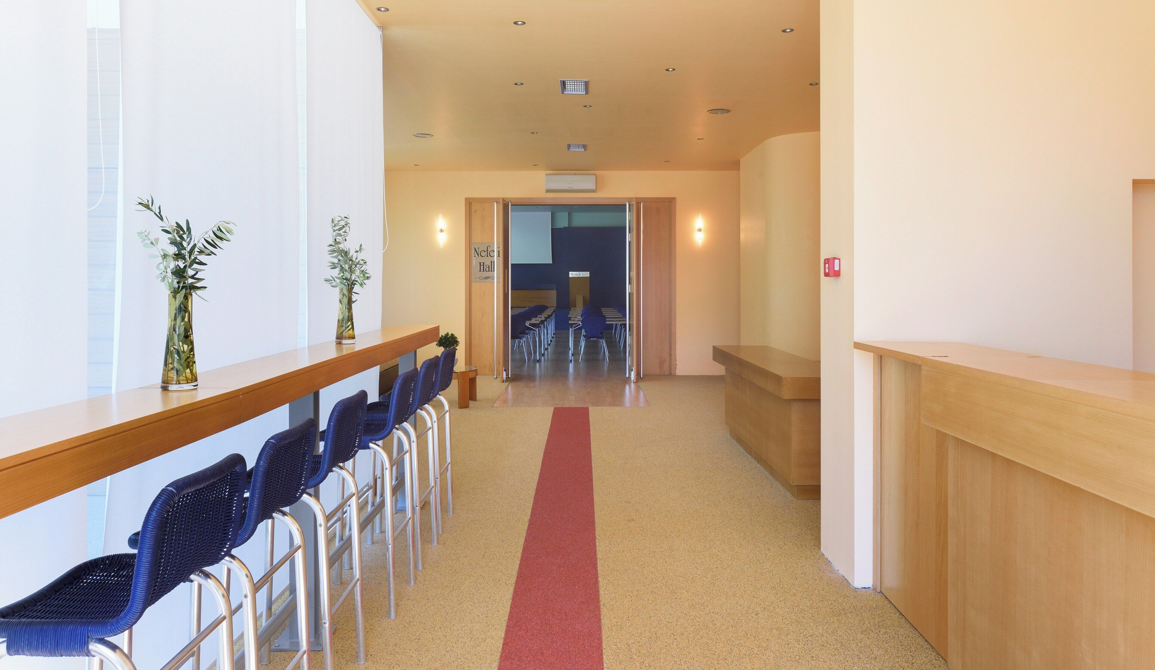 Un hall de hotel con mostrador de recepción, zona de estar con sillas, alfombra roja y un cartel para el Nefeli Hall.