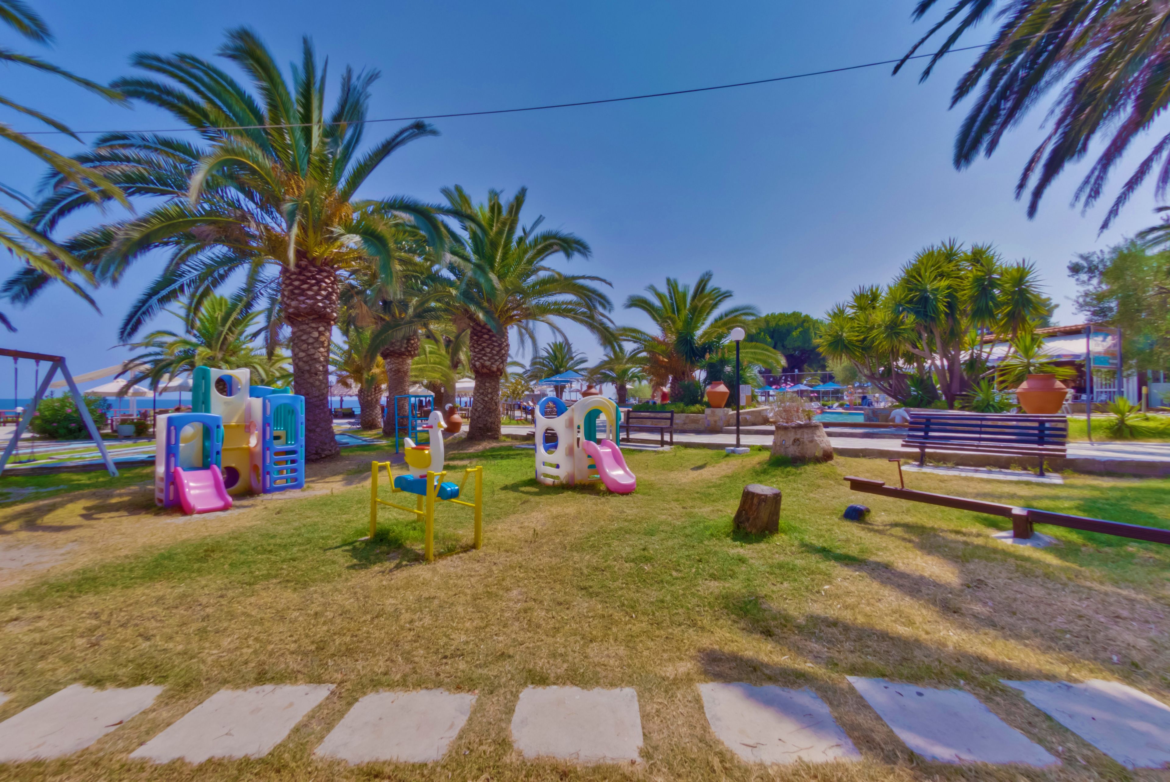 A playground with slides and climbing structures, surrounded by palm trees and benches.